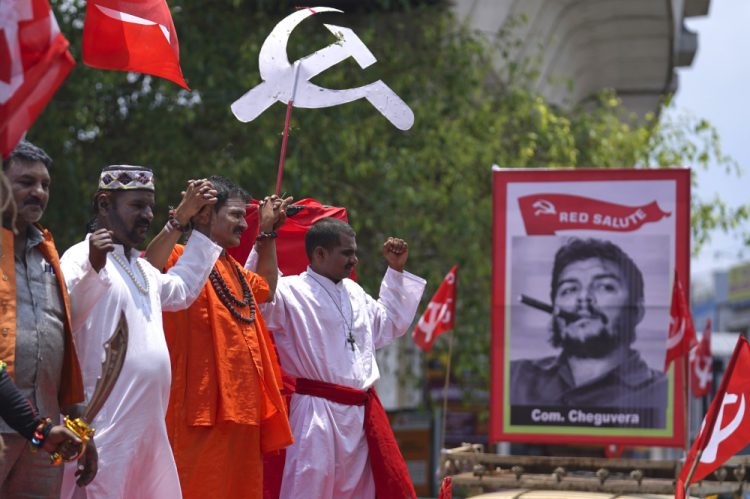 Členovia Centra indických odborov (CITU) oblečení ako moslimskí, hinduistickí a kresťanskí kňazi stoja vedľa obrazu kubánskeho revolučného vodcu Che Guevaru 1. mája 2023 (Foto: SITA/AP Photo/Mahesh Kumar A.)