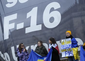 Protestujúci, niektorí zabalení v ukrajinských vlajkách, stoja vedľa nápisu žiadajúceho amerického prezidenta Joea Bidena, aby poslal stíhačky na Ukrajinu v Poľsku v stredu 22. februára 2023 (Foto: SITA/AP Photo/Alastair Grant)